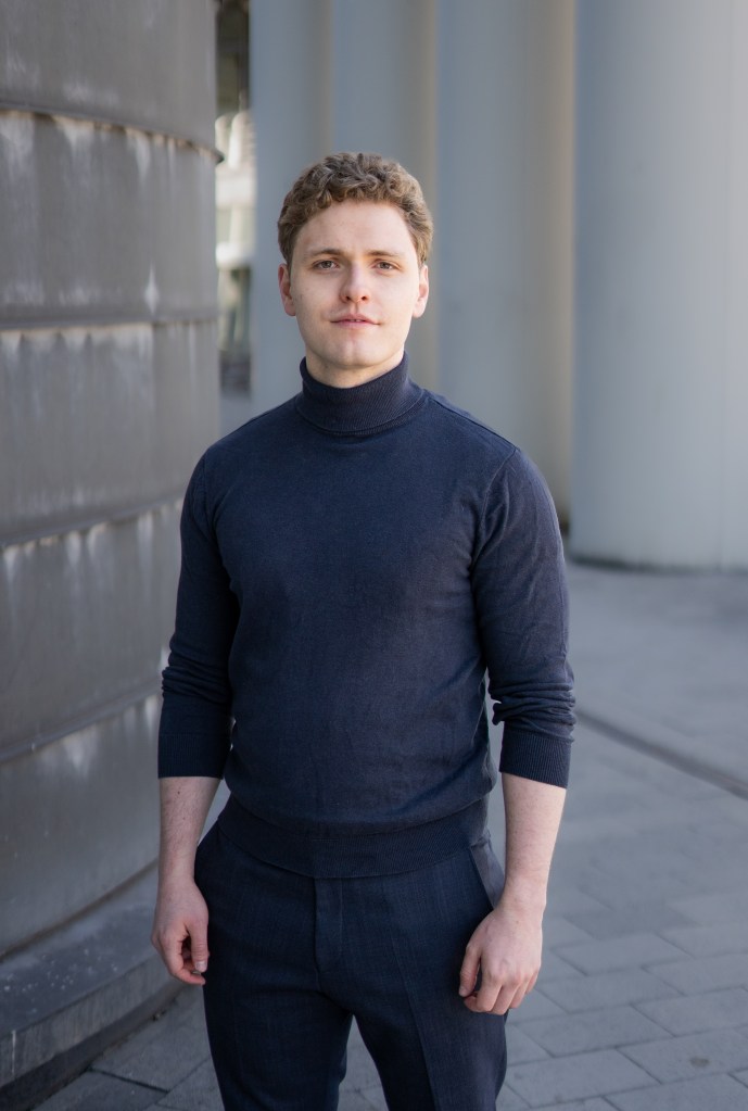 Lukas Baeskow steht selbstbewusst in Rollkragen im Ganzkörperbild