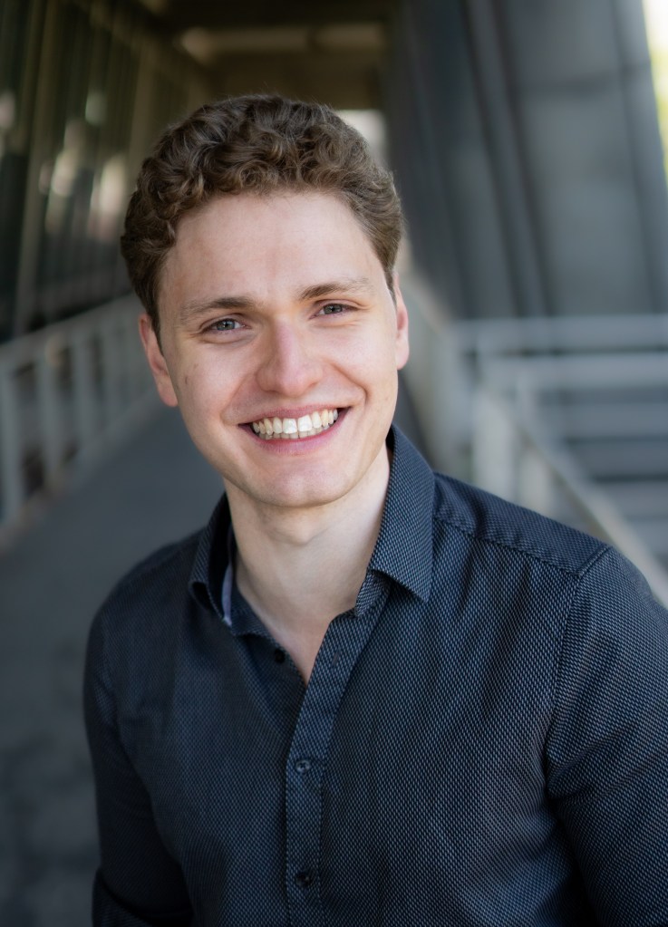 Lukas Baeskow lächelt natürlich und zeigt seine Zähne in grauem Hemd