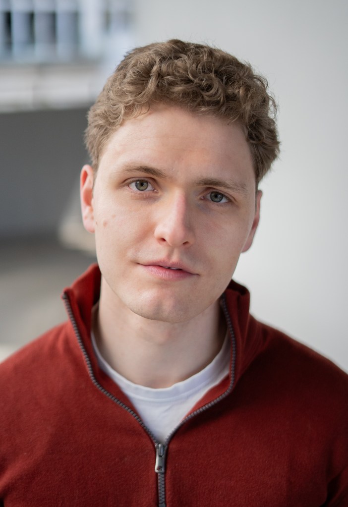 Lukas Baeskow schaut mit neutralem und ernstem Blick in rotem Pullover